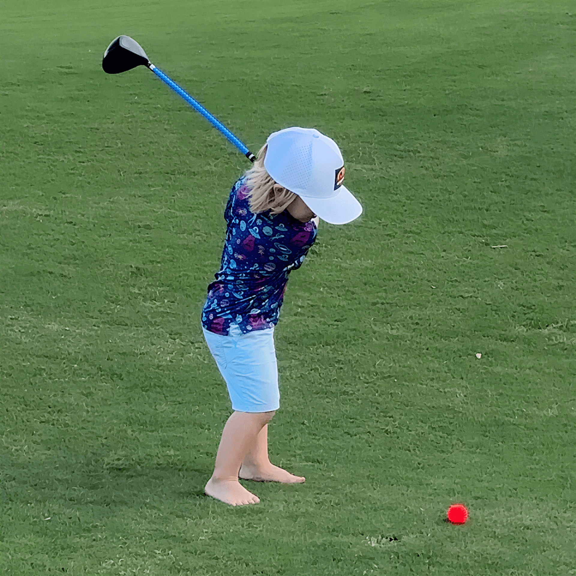A young child in a white hat and patterned shirt swings a blue golf club at a red ball on a green grass field.